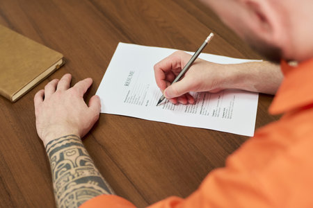 Caucasian young adult man with tattooed arm sitting at table filling out resume form with pencil, focusing on handwriting, education in prison context, brown notebook nearbyの写真素材