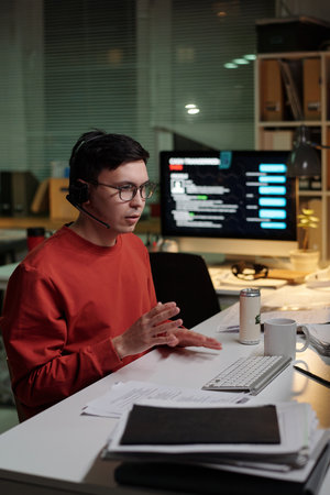 Young adult man wearing headset sitting at desk operating computer, engaging in scam call center activity during nighttime shiftの写真素材