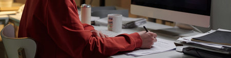 Man sitting at desk using computer and writing on documents with pen, working in scam call center environment with paperwork and office suppliesの写真素材