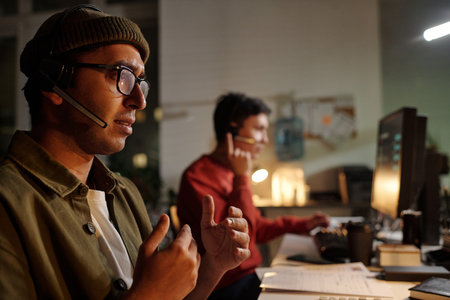 Indian man wearing headset operating computer and gesturing while sitting beside young adult man...