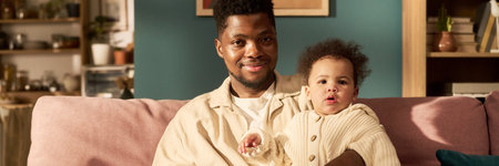 Black young adult man sitting on sofa holding infant child, both looking toward camera, father...