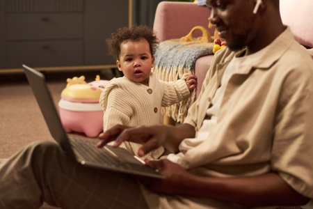 Black young adult man sitting on floor using laptop while caring for baby standing nearby, baby...
