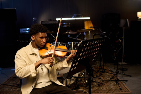 Young adult Black man playing violin in music studio, sitting behind music stand with focused...