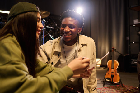 Young Black man smiling and interacting with young Hispanic woman holding paper in music studio,...