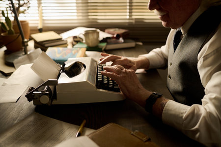Senior Caucasian man typing on vintage typewriter at desk, hands on keys, wearing formal vest, surrounded by papers and books, sunlight streaming through window blindsの写真素材