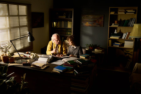 Senior Caucasian man and middle aged Caucasian woman sitting at desk discussing documents surrounded...