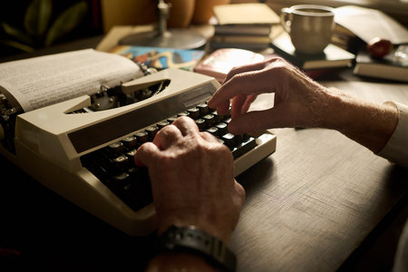 Senior Caucasian man typing on vintage typewriter with both hands, working at wooden desk with open book and coffee cup visible in background, focused on writing processの写真素材