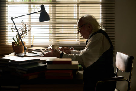 Senior Caucasian man sitting at desk writing on paper, surrounded by stacked books and office supplies, concentrating on work in front of window with blinds partially closedの写真素材