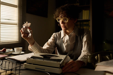 Caucasian middle aged woman sitting at desk holding crumpled paper in hand, wearing glasses, looking...