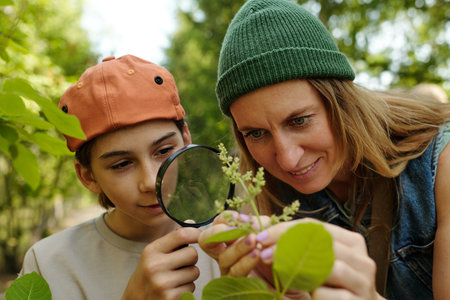 Caucasian woman and Caucasian teenager examining plant leaves with magnifying glass outdoors, both focusing intently on details of foliage, engaging in nature exploration togetherの写真素材