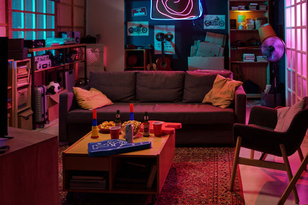 Modern living room featuring empty gray sofa and armchair, illuminated by neon lights, coffee table holding snacks, drinks, party cups, and tabletop game, shelves with books and guitars in backgroundの写真素材