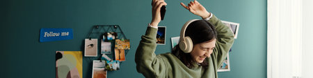 Teenage girl listening to music with headphones, smiling and raising arms while holding smartphone,...