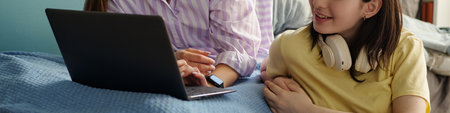 Girl sitting on bed using laptop with teenage friend wearing headphones, both smiling and...