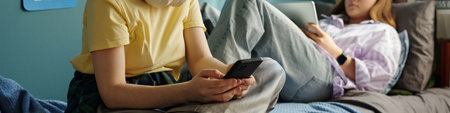 Two Caucasian teenagers using digital devices in bedroom, one sitting on bed holding smartphone,...