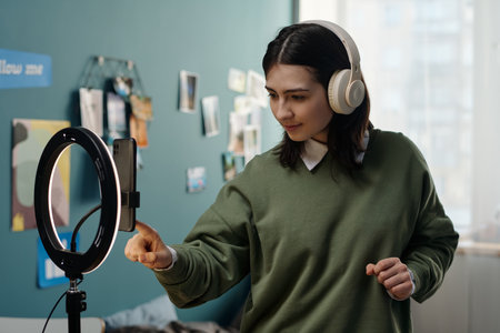 Young adult Caucasian woman wearing headphones interacting with smartphone mounted on ring light tripod in bedroom, recording video content or livestreaming for social media platformの写真素材