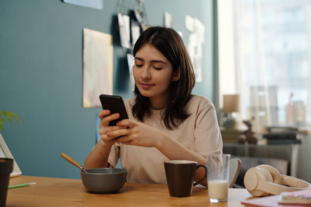 Young adult Caucasian woman sitting at table using smartphone while eating breakfast, holding phone with both hands, bowl and cup on table, headphones and glass of milk nearbyの写真素材