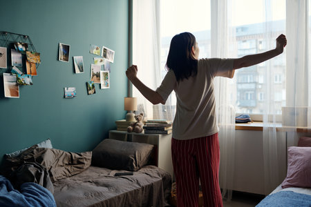 Young adult Caucasian woman standing in bedroom stretching arms while looking out window in morning, sunlight streaming in, casual home environment with personal photos on wallの写真素材