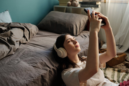Teenage Caucasian girl lying on floor next to bed listening to music with over ear headphones, holding smartphone above face relaxing in bedroom during daytimeの写真素材