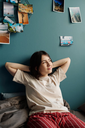 Teenage Caucasian girl woman relaxing on bed with hands behind head, gazing thoughtfully to side,...