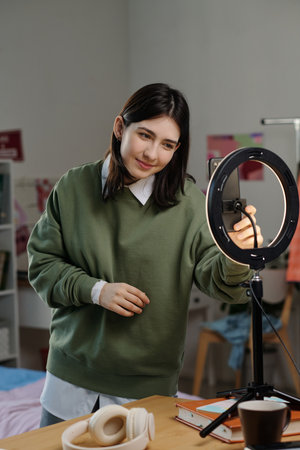 Young adult Caucasian woman adjusting smartphone on ring light tripod in home setting, preparing for video recording or live streaming, focused expression, casual postureの写真素材
