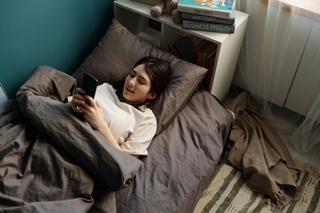 Teenage Caucasian girl lying in bed using smartphone, smiling while holding device with both hands,...