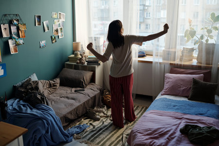 Young adult Caucasian woman standing near window stretching arms in shared bedroom with two beds, morning light coming through sheer curtains, personal items scattered on floorの写真素材