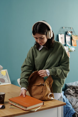Teenage Caucasian girl wearing headphones packing brown backpack with books and notebooks standing...