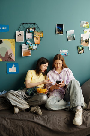 Two teenage girls sitting on bed sharing potato chips while looking at smartphone together, wall...