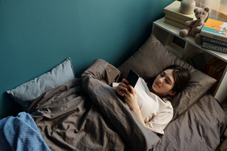 Teenage Caucasian girl lying in bed using smartphone, resting against pillows with books and teddy...