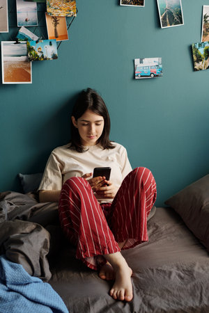 Caucasian teenage girl sitting on bed using smartphone, relaxing against teal wall decorated with...