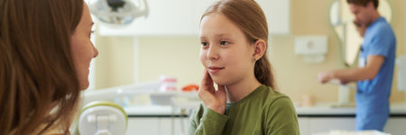 Caucasian girl touching cheek while talking to Caucasian woman in dental clinic, male dentist...
