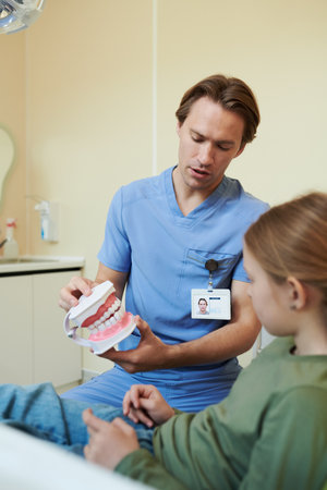 Caucasian young adult man dentist demonstrating dental hygiene using teeth model while sitting with Caucasian child girl patient in dental clinic, both focusing on modelの写真素材