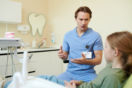 Caucasian young adult male dentist explaining dental procedure to Caucasian child girl patient sitting in dental chair, dental equipment and model teeth visible in clinic backgroundの写真素材