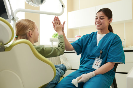Caucasian child sitting in dental chair giving high five to young adult woman dentist wearing scrubs and gloves, both smiling and interacting in dental office settingの写真素材