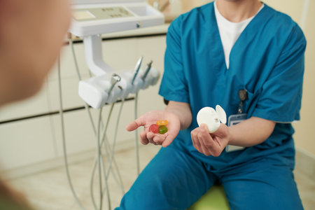 Caucasian young adult woman wearing medical scrubs sitting in dental office holding dental floss container in one hand and colorful gummy candies in other hand, while talking to patientの写真素材