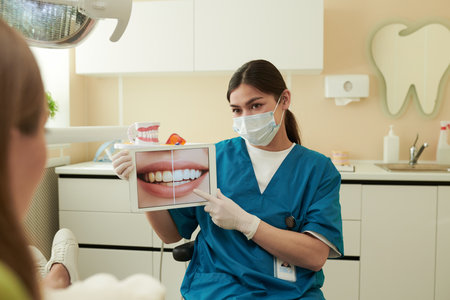Young adult Caucasian woman dentist wearing medical mask and gloves sitting in dental clinic, showing patient dental treatment results on digital tablet displaying healthy teethの写真素材
