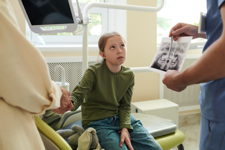 Caucasian girl child sitting on dental chair looking at dentist while dental professional showing dental X-ray to adult woman, medical consultation in dental clinicの写真素材