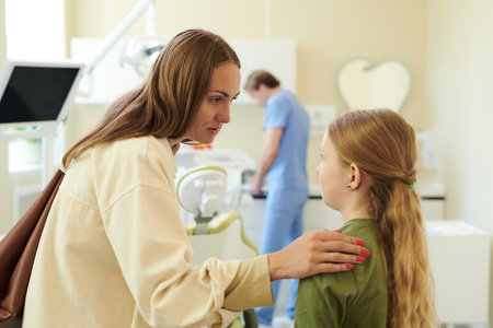 Caucasian woman comforting Caucasian girl with hand on shoulder in dental clinic while medical professional working in background, both subjects facing each other, girl looking attentiveの写真素材