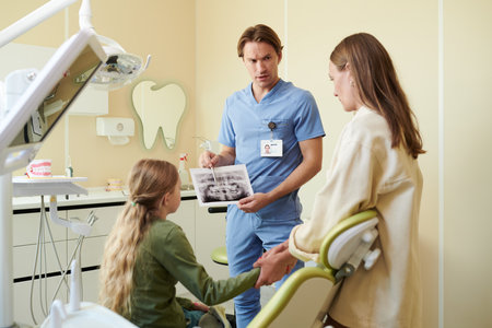 Caucasian male dentist showing dental X-ray to Caucasian teenage girl sitting in dental chair while Caucasian woman, likely mother, standing beside holding teenagers handの写真素材