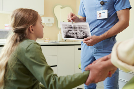 Caucasian preteen girl sitting in dental office listening to male dentist explaining dental X-ray while holding adult hand, dentist wearing scrubs and showing radiographの写真素材
