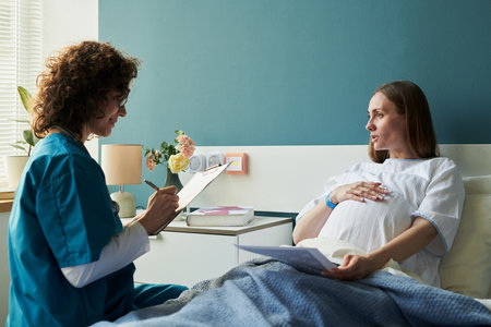 Caucasian pregnant young adult woman sitting in hospital bed talking with Caucasian female nurse holding clipboard, nurse writing notes while patient touching belly, medical consultation sceneの写真素材