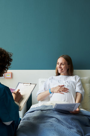 Caucasian pregnant young adult woman lying in hospital bed smiling and holding ultrasound image...