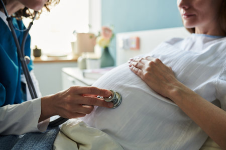Caucasian young adult woman lying in hospital bed receiving prenatal checkup from female healthcare professional using stethoscope on pregnant belly during medical examinationの写真素材