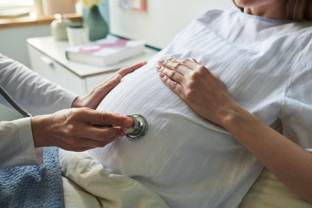 Caucasian pregnant woman lying on hospital bed receiving prenatal checkup from medical professional using stethoscope on abdomen, hand resting gently on bellyの写真素材