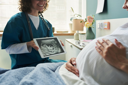 Caucasian pregnant woman lying on examination table holding belly while female medical professional showing ultrasound image on digital tablet during prenatal checkupの写真素材