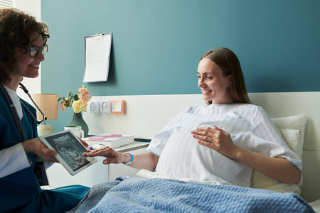 Caucasian pregnant young adult woman lying in hospital bed smiling at female doctor showing ultrasound image on digital tablet during prenatal checkup in medical clinicの写真素材