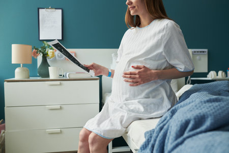 Pregnant Caucasian young adult woman sitting on hospital bed holding ultrasound image, gently touching belly with one hand, wearing medical gown, smiling slightly, side viewの写真素材