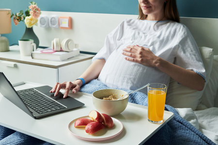 Pregnant Caucasian woman sitting in hospital bed using laptop with left hand while holding belly with right hand, breakfast tray with oatmeal, apple slices, orange juice in foregroundの写真素材
