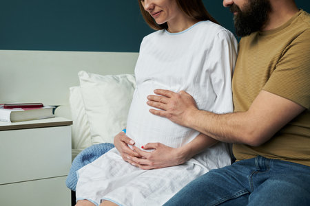 Caucasian pregnant woman sitting on bed holding belly with middle aged Caucasian man gently touching her stomach, both looking down and sharing intimate moment in hospital roomの写真素材
