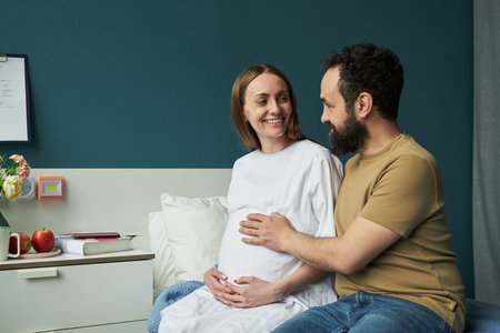 Caucasian pregnant young woman sitting on bed smiling while Caucasian young man touching her belly, both looking at each other in bedroom setting with books and fruit on nightstandの写真素材